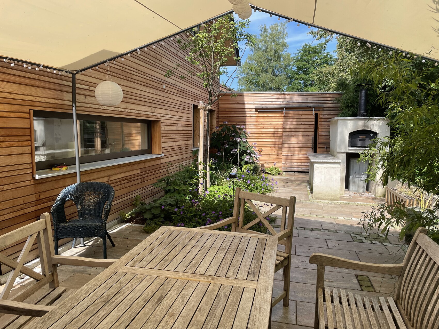 Terrassenbereich vor der Holzhaus-Wand mit Esstisch, Stühlen, Korbsessel und Grill rechts.