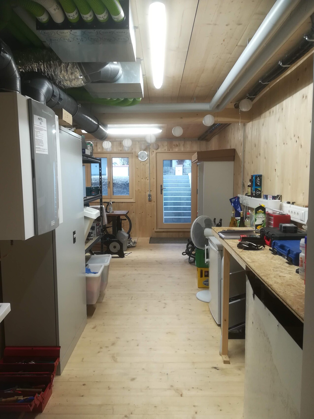 Holzwerkstatt mit Deckenrohren, rechts Werkbank mit Werkzeug, links Schrank, Tür führt nach außen.