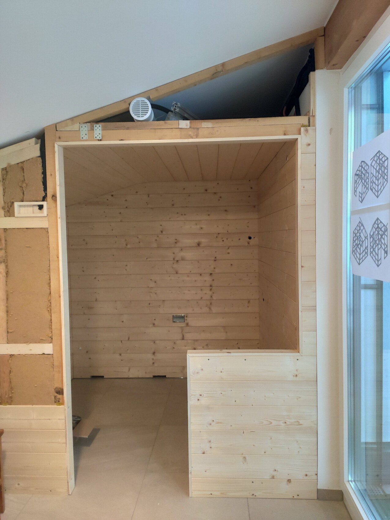 Heller Innenraum aus Holzlatten, rechteckiger Raum mit offener Tür, sichtbare Dachkonstruktion.