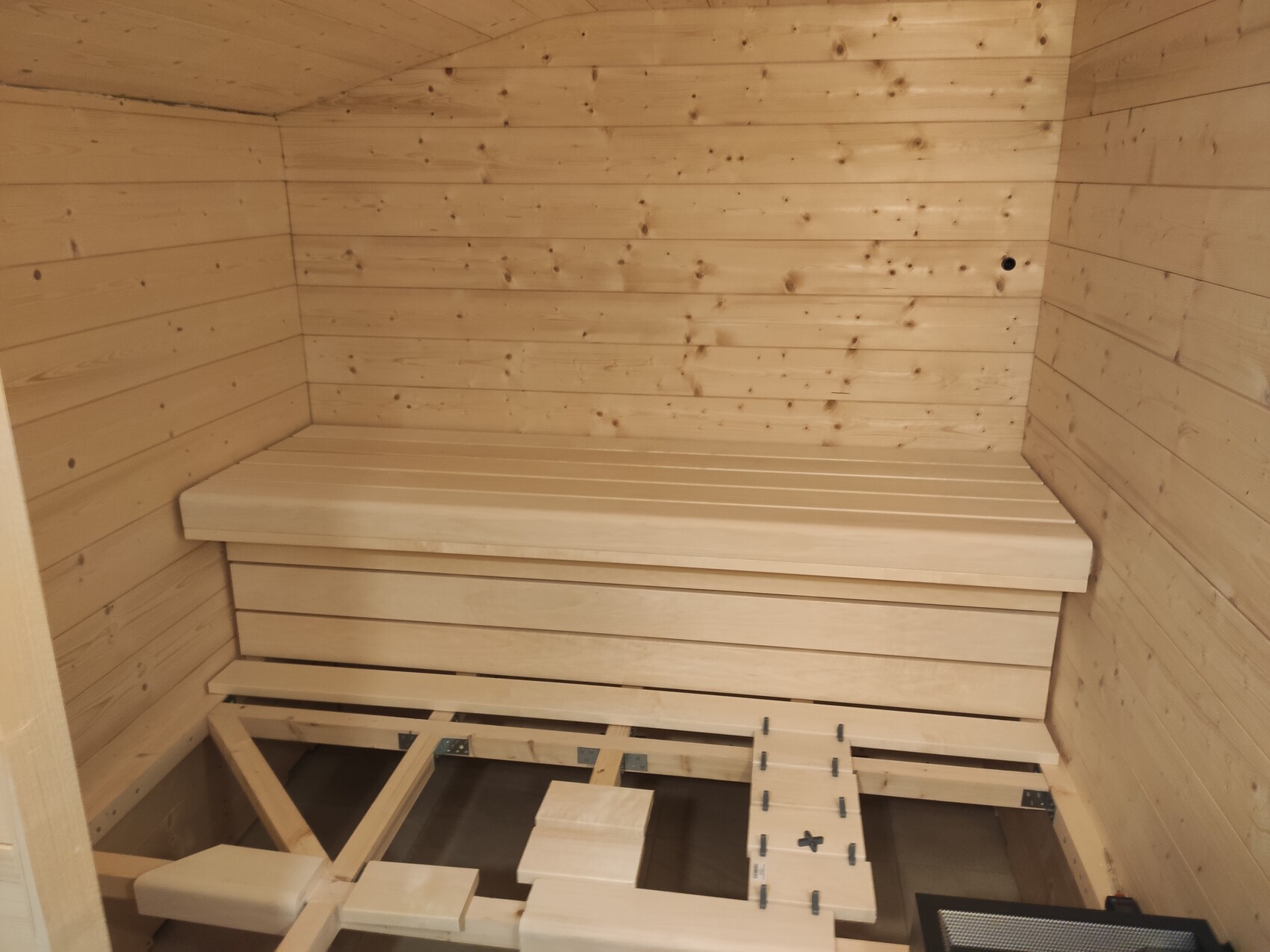 Holzsauna-Innenraum mit Bank, Holzwänden und Heizofen unten rechts.