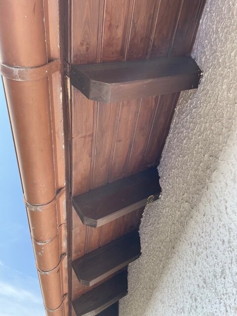 Holzwand mit drei herausstehenden Brettern als Stufen; links braunes Regenrohr, rechts Putzwand.