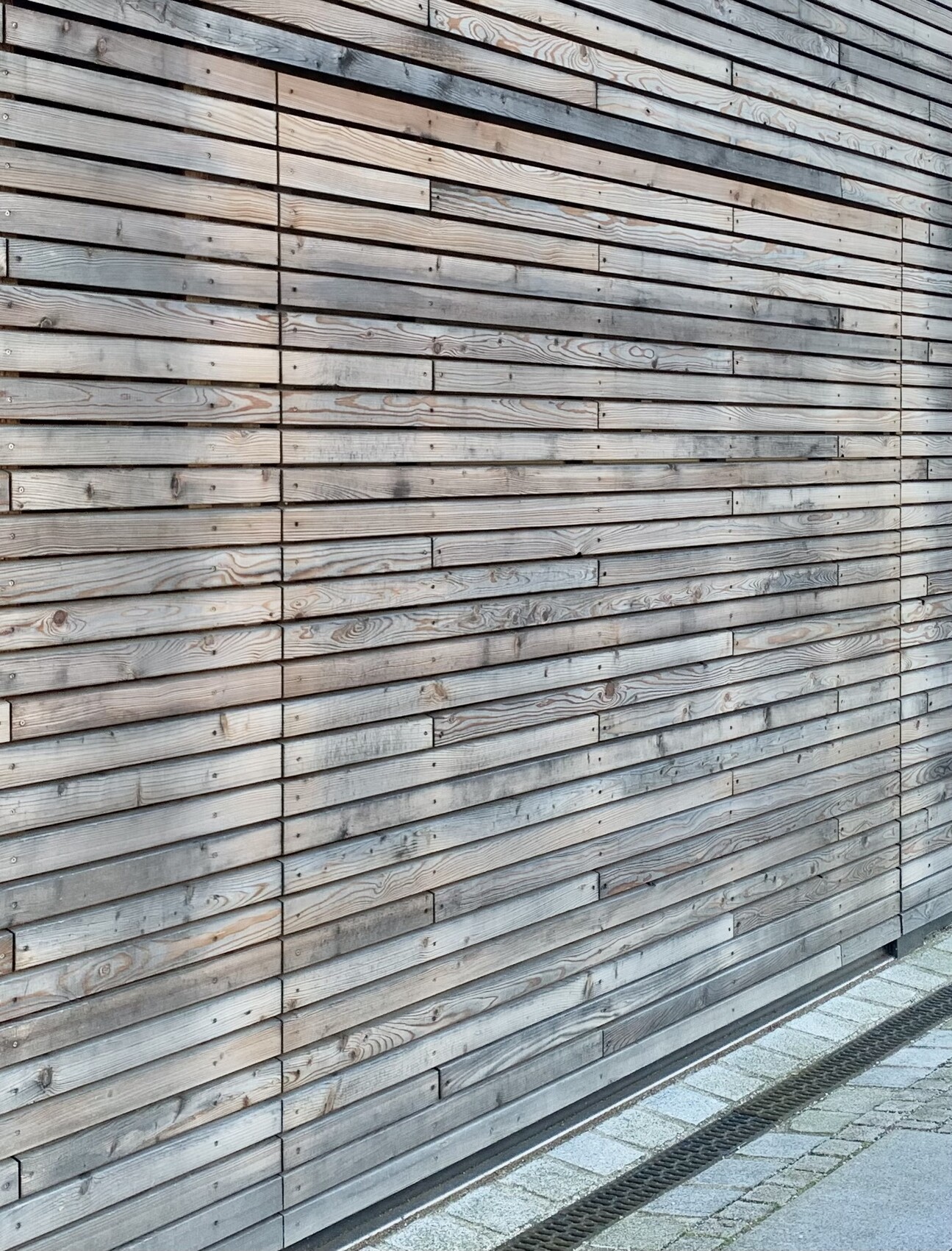 Horizontale verwitterte Holzbretter-Wand vor einem gepflasterten Gehweg mit einer Abflussrinne.
