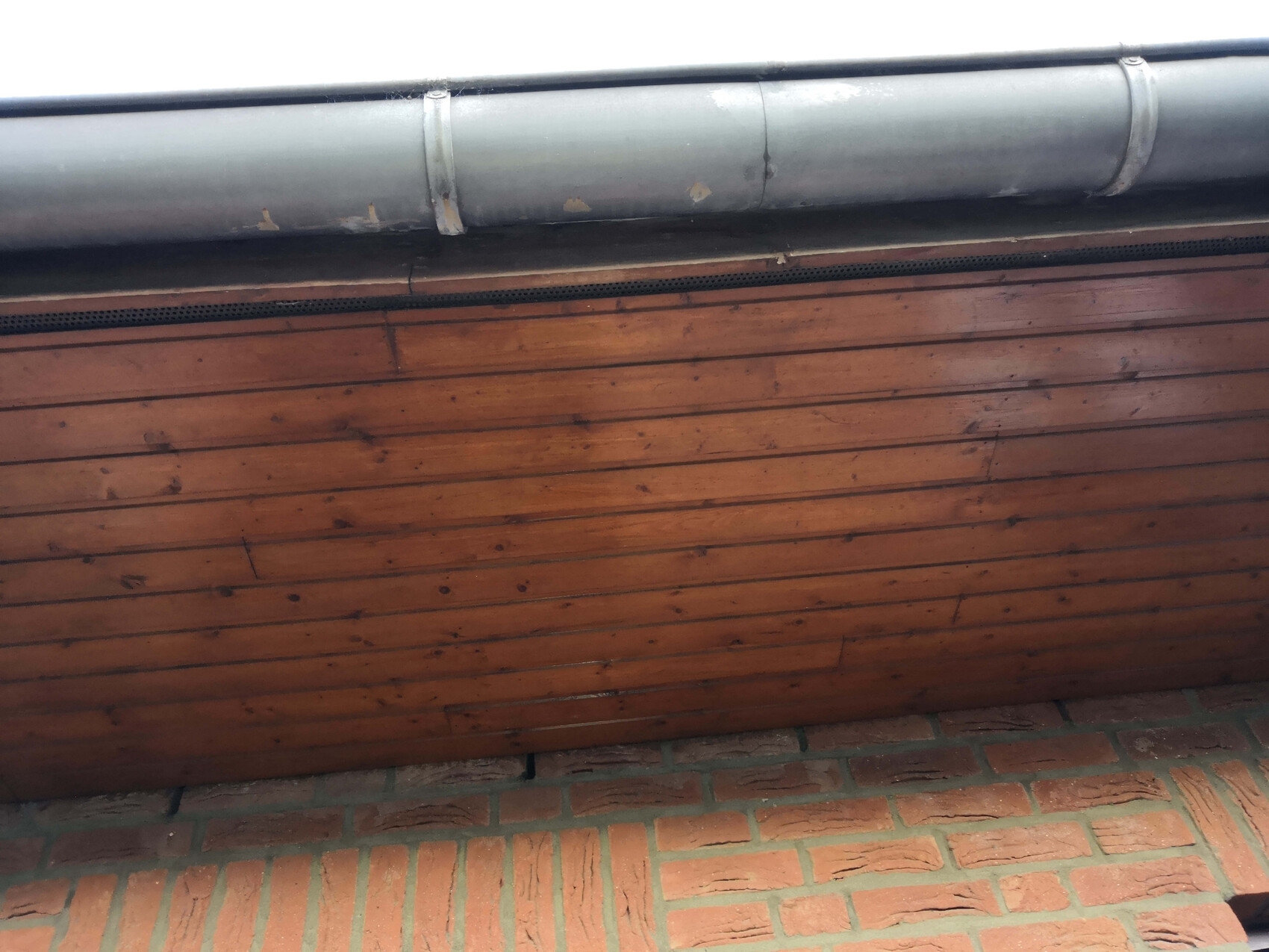 Holzwand mit horizontalen Brettern, darüber graues Rohrsystem, darunter Ziegelboden.