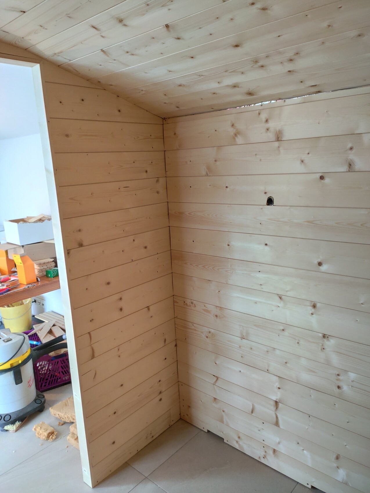 Winkel eines Raums mit hellen Holzpaneelen an Wänden und Decke; Werkstattbereich im Hintergrund.
