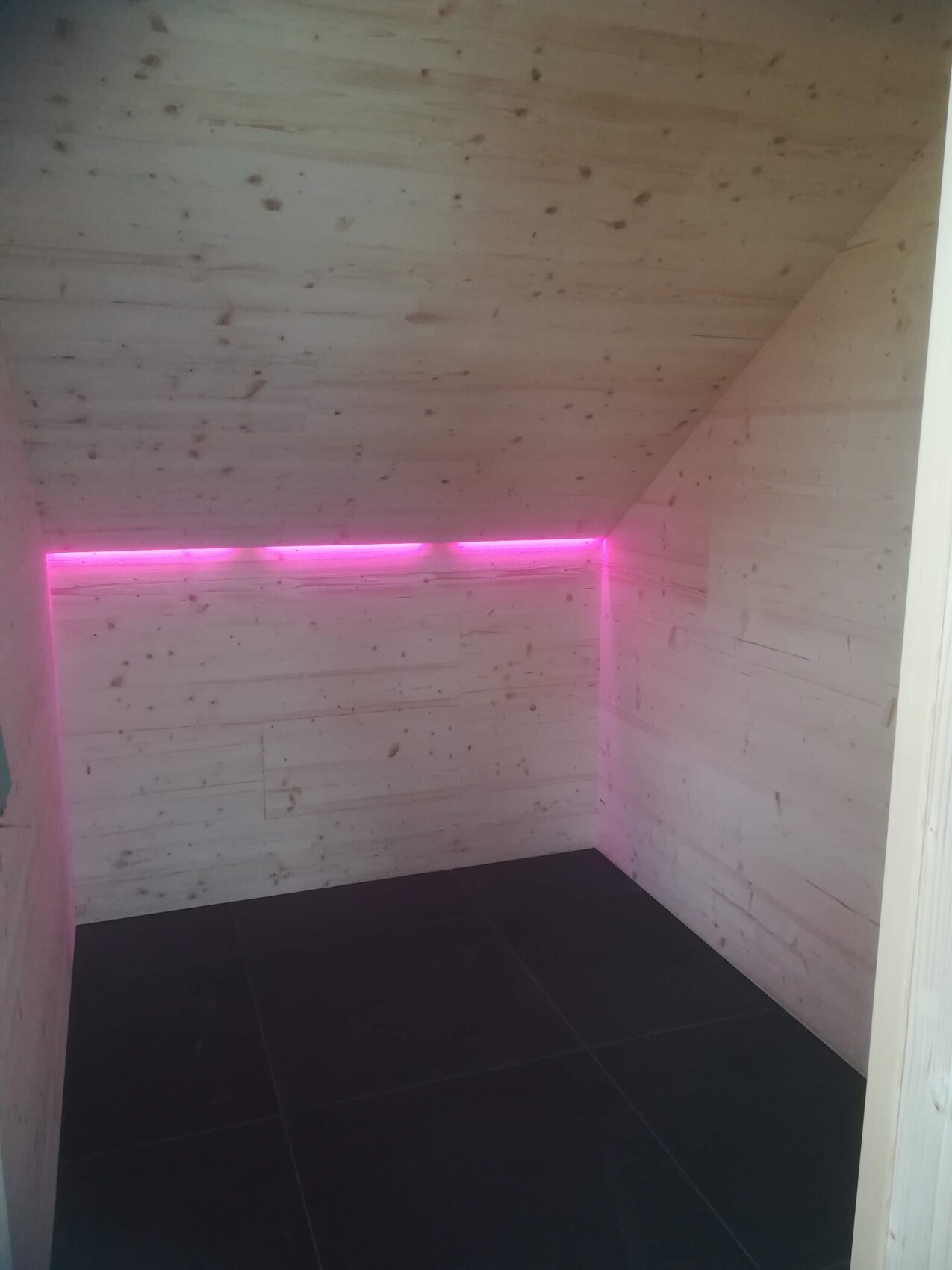 Kleiner Raum mit Holzverkleidung, dunklem Boden, pinkem LED-Licht an der Wand und schräge Decke.