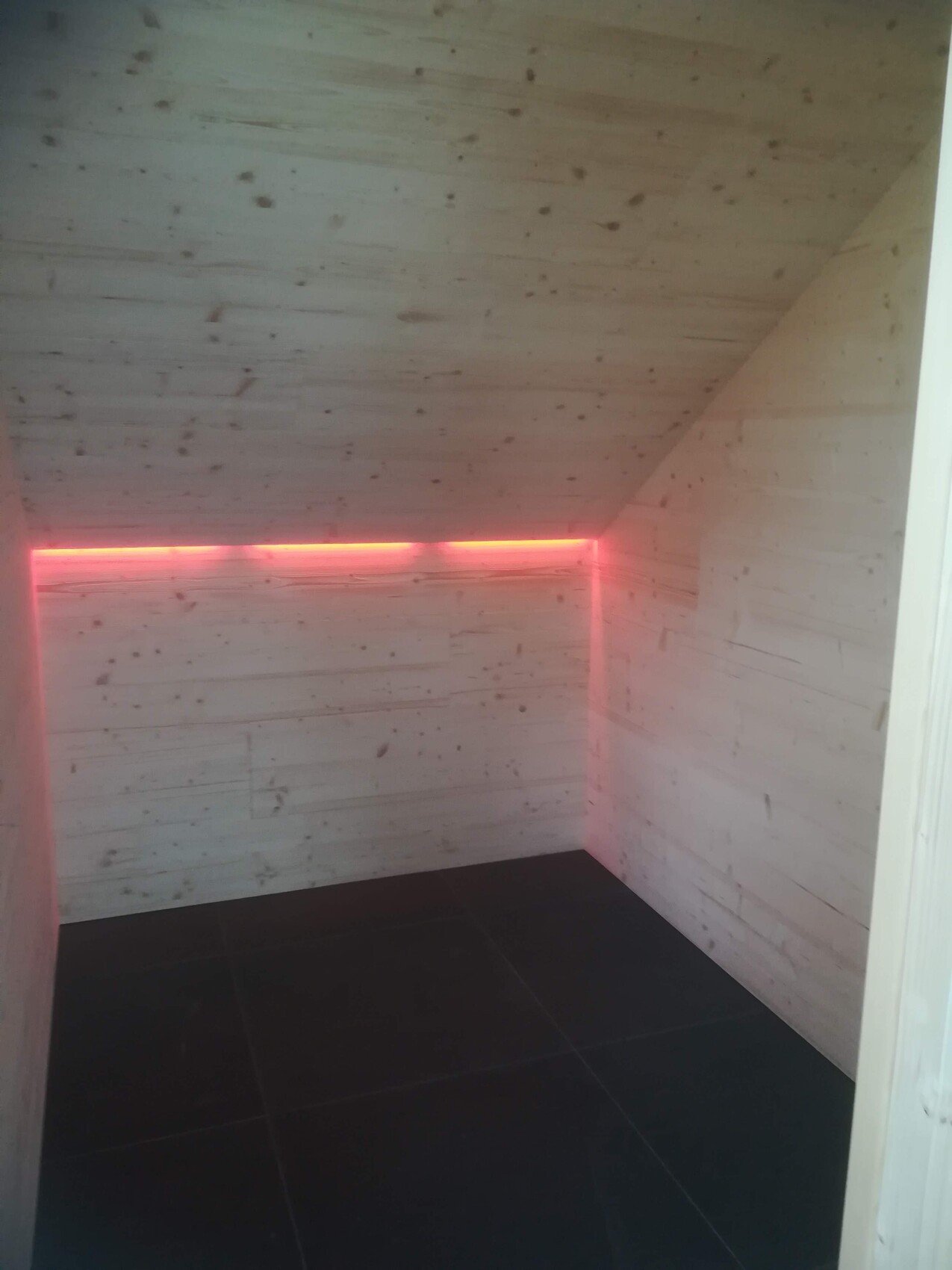 Kleiner Holzraum mit schrägem Dach, dunklem Fliesenboden und pinkroter LED-Lichtkante in der Ecke.