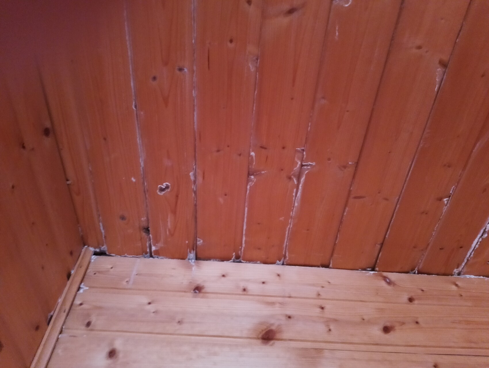 Ecke eines Raums mit vertikalen Holzwänden und Holzboden; weiße Fugenabdichtung sichtbar.
