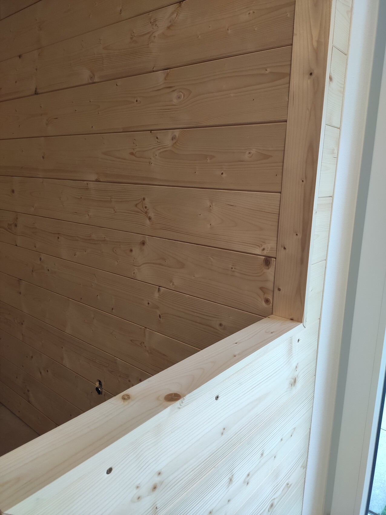 Heller Innenraum mit Holzwand aus horizontalen Brettern und sichtbarem Fensterrahmen rechts.