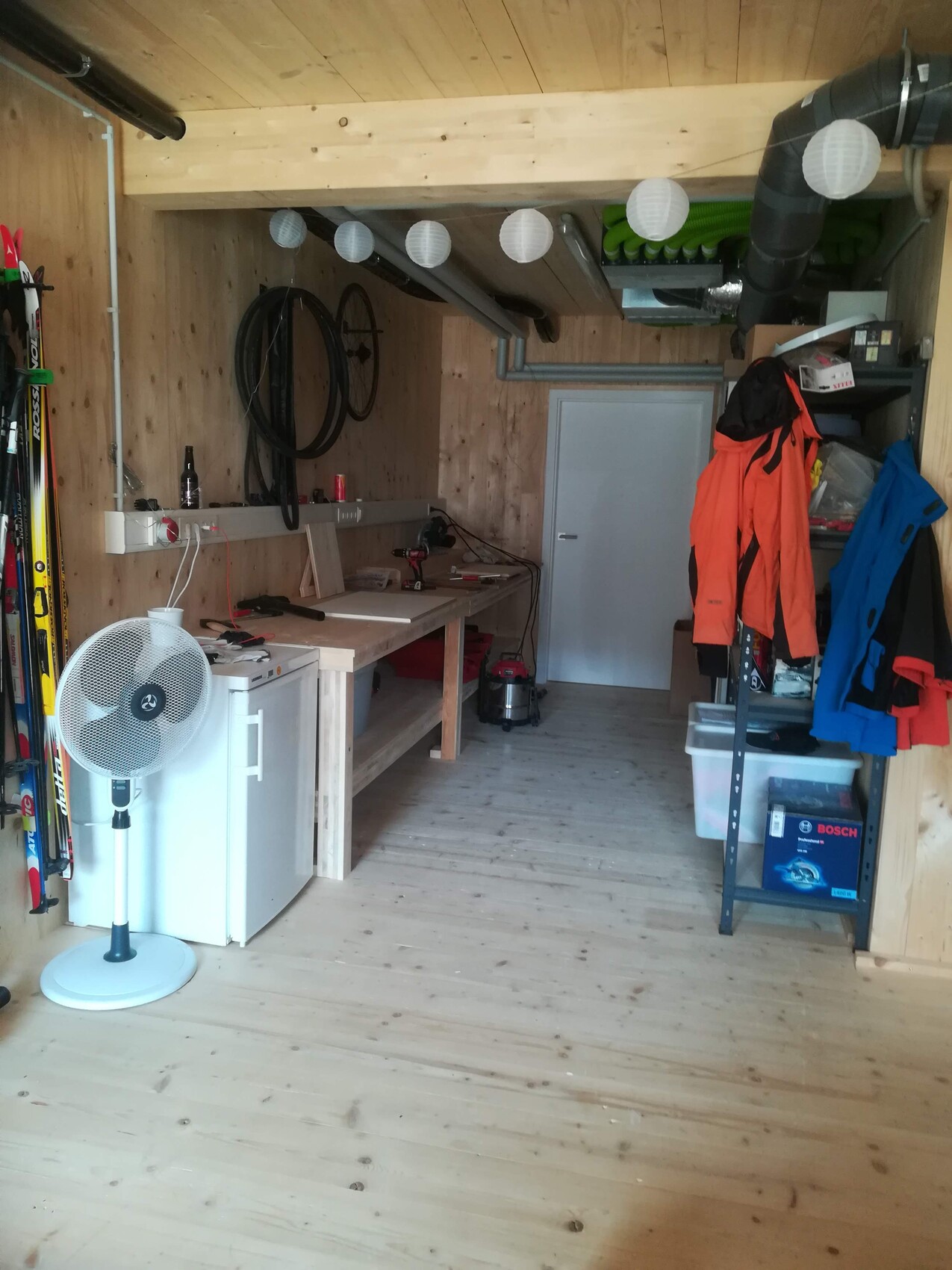 Holzwerkstatt mit Arbeitsbank, Fahrradräder an Wand, Standventilator, Jacken am Regal