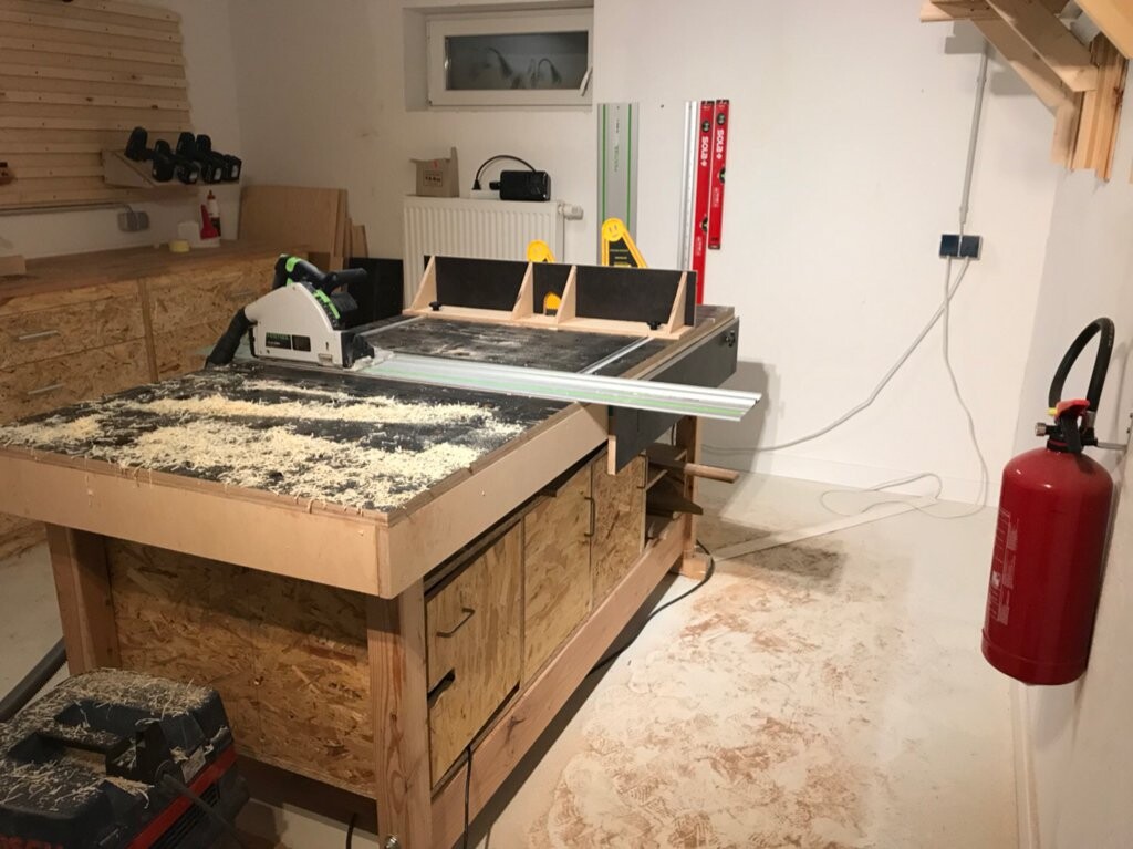 Werkstatt mit Tischsäge auf einer Holzwerkbank, Späne und Holzreste, Feuerlöscher an der Wand.