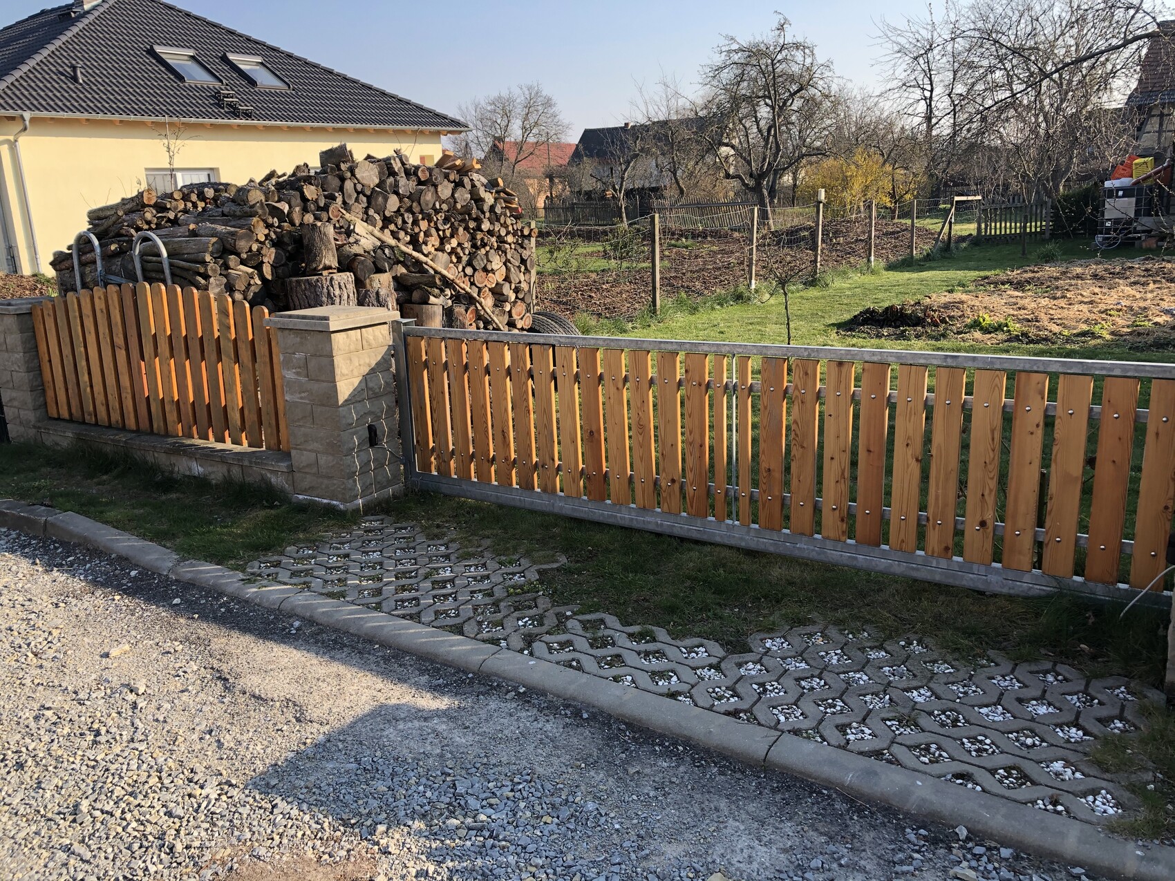 Gelbes Haus links, großer Holzstapel hinter Holzlattenzaun, gepflasterter Weg im Vordergrund.