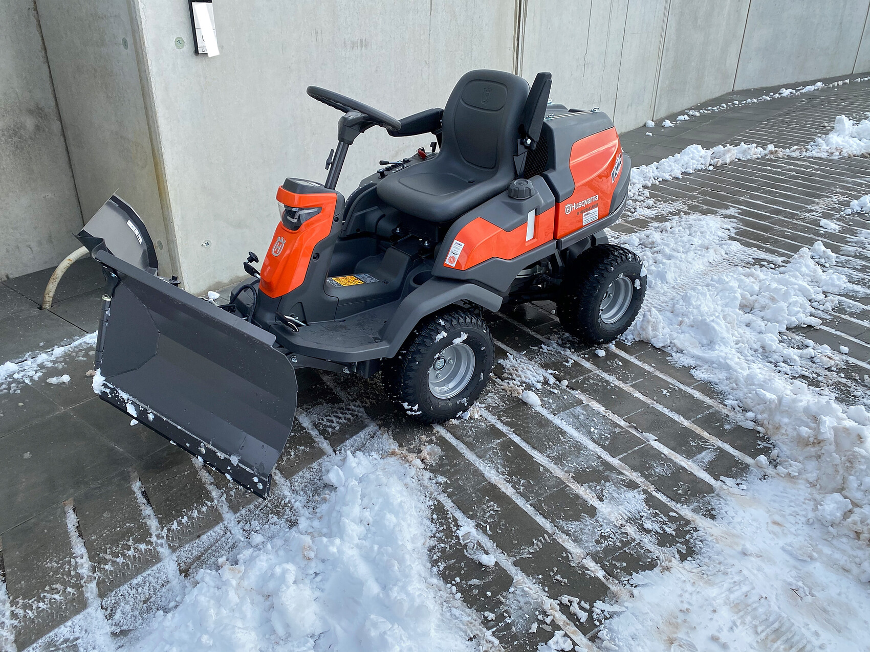 Kleiner orange-grauer Husqvarna-Schneepflug mit Sitz und Frontschaufel im Schnee.