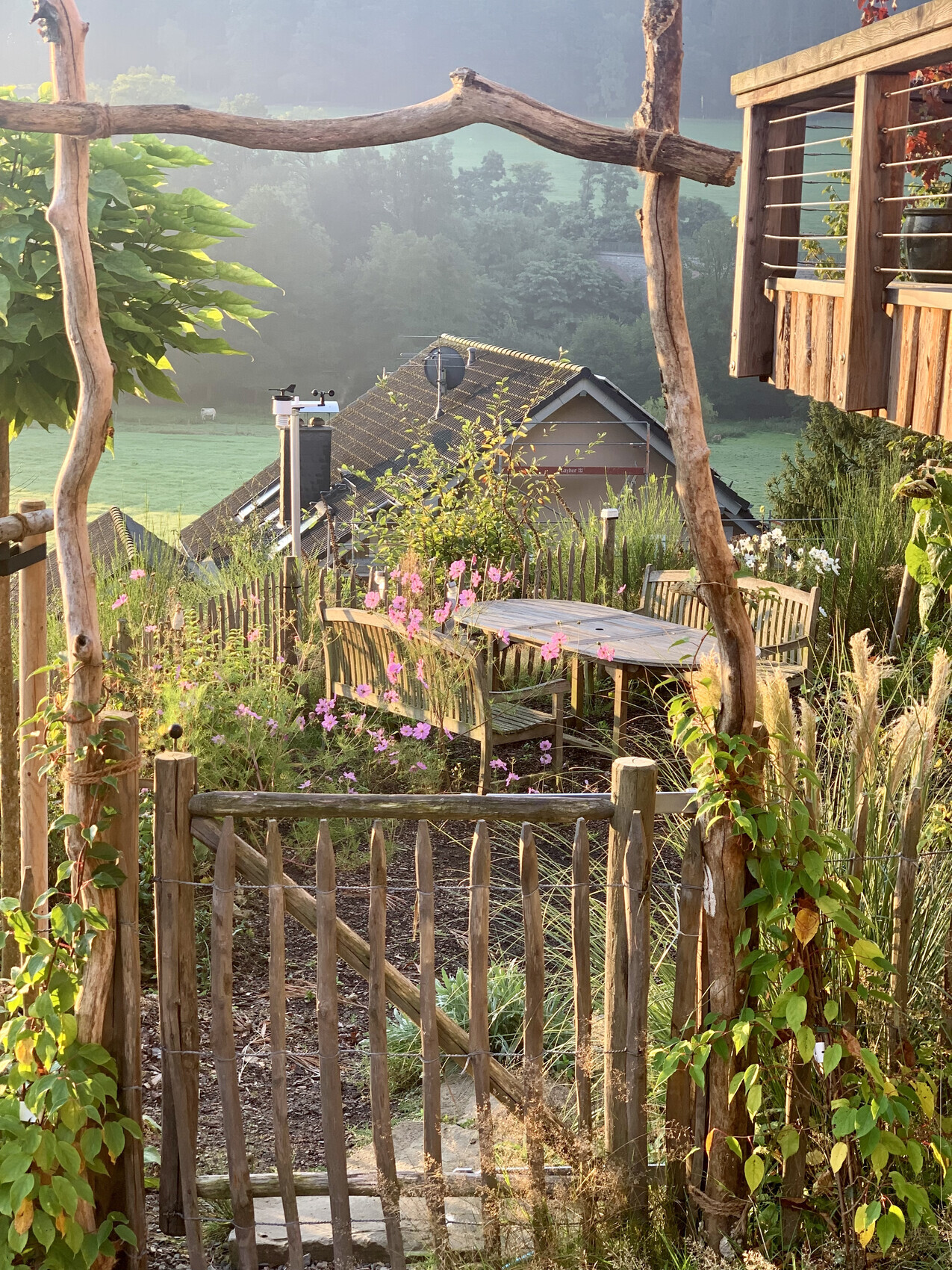 Rustikale Gartenidylle: Holzzaun, Tischgruppe, Blüten, Blick auf Dach und grüne Landschaft