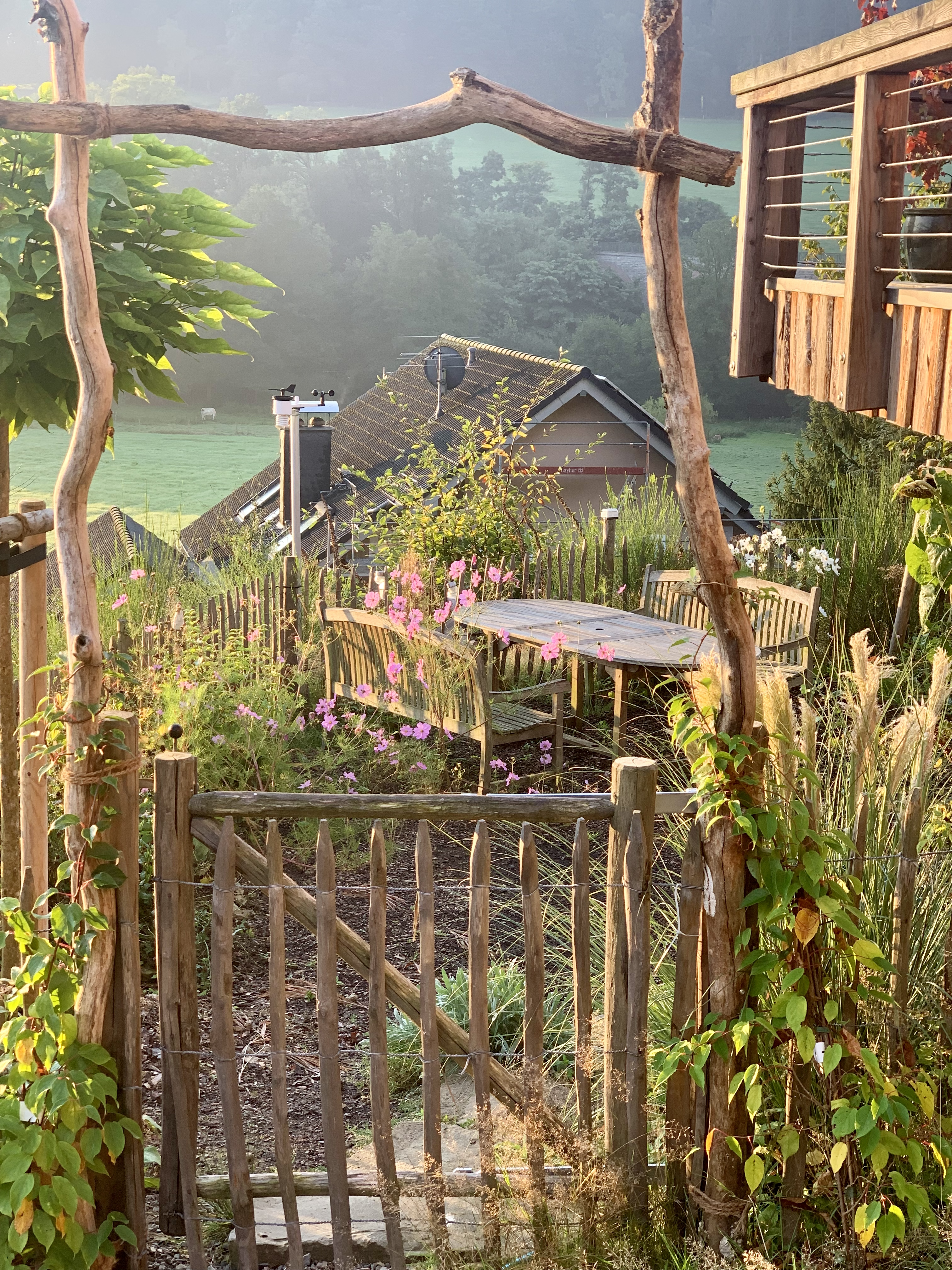 idyllische-garten-terasse-mit-holzmoebel-und-blumen-am-hang.png