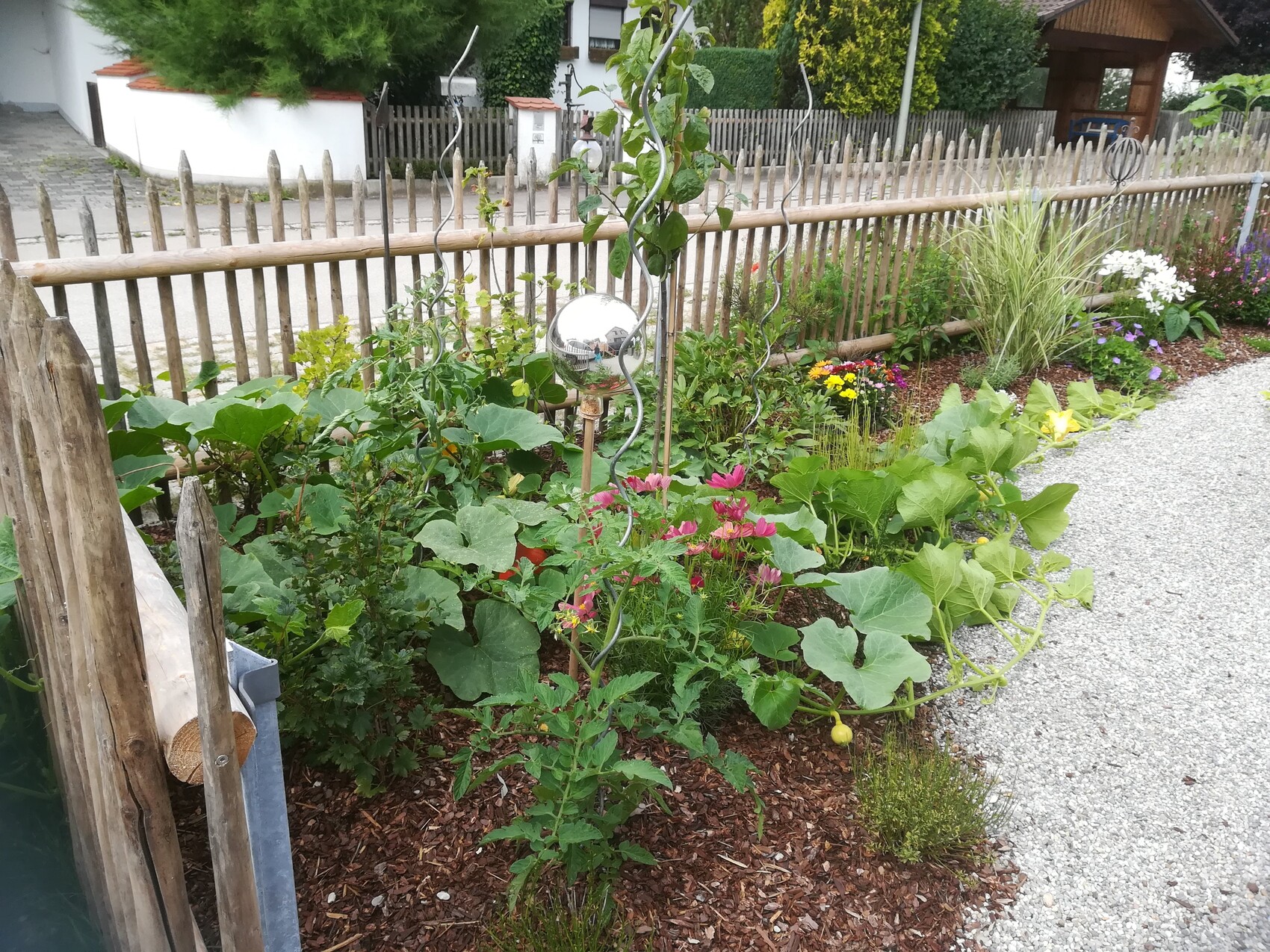 Gartenbeet am Holzzaun mit grünen Blättern, bunten Blumen und silberner Kugel auf einer Spirale