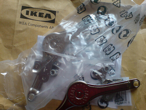 IKEA-Verpackung mit Metallteilen, Plastiktüte und rotem Arm/Klemmteil.