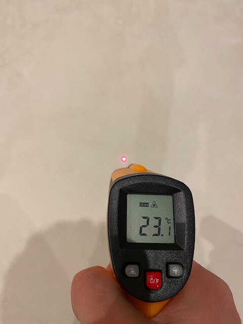 Hand hält ein Infrarot-Thermometer; Display zeigt 23,1 °C, roter Laser zielt auf Wand.