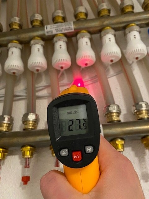 Hand hält IR-Thermometer vor Rohrleitungen mit Ventilen; Anzeige 21,5°C.