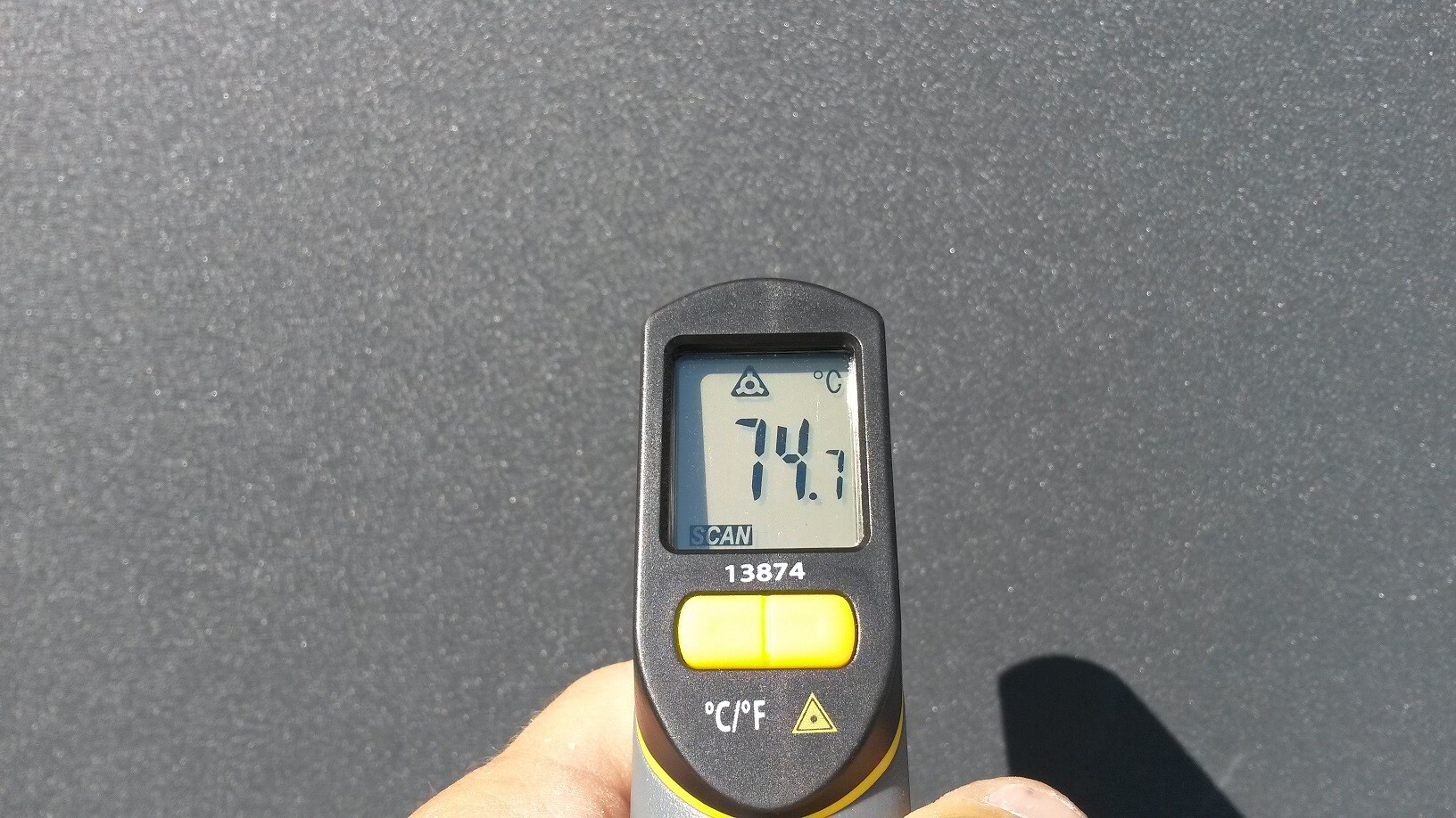 Handheld Infrarot-Thermometer zeigt 74,7 °C auf dunklem Asphalt