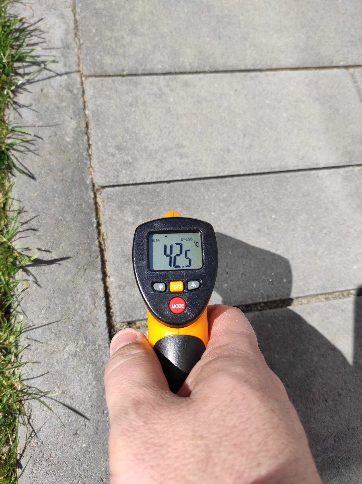 Hand hält Infrarot-Thermometer über Gehwegplatten; Display zeigt 42.5°C