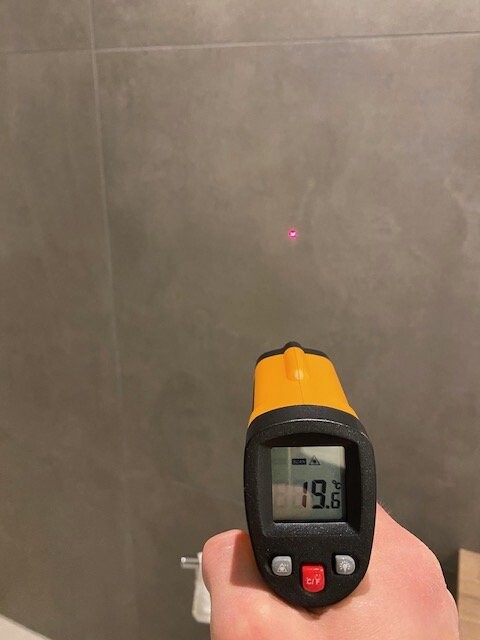 Handgehaltenes Infrarot-Thermometer zeigt 19,6 °C; roter Laserpunkt auf grauer Wand.