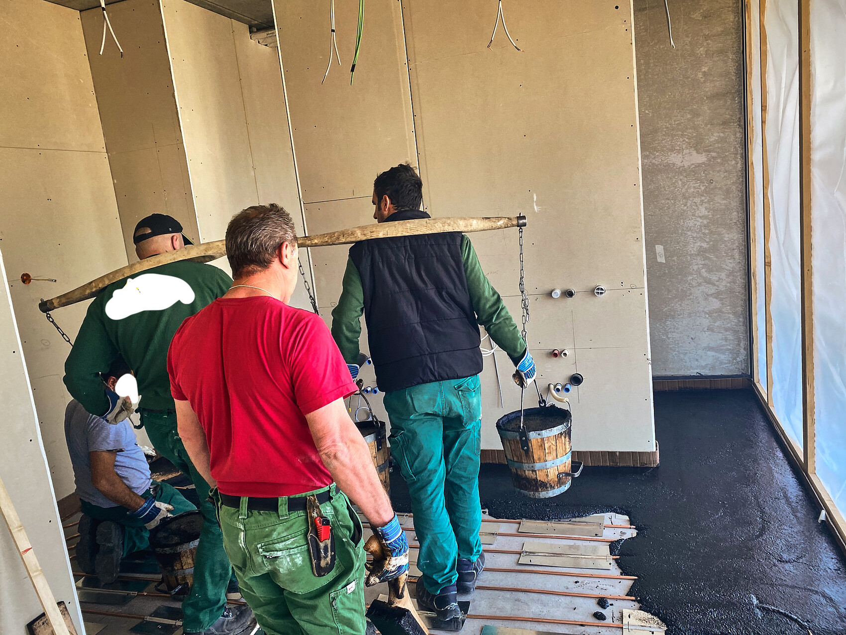 Mehrere Bauarbeiter tragen einen Eimer Beton an einer langen Holzstange im Innenausbau.