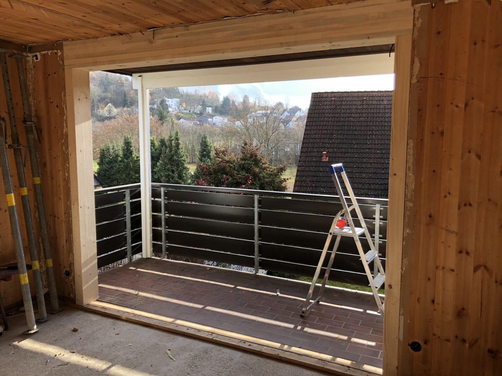 Rohbauzimmer mit offener Balkontür, Balkon mit Leiter und Brüstung, Blick auf Dächer und Bäume.