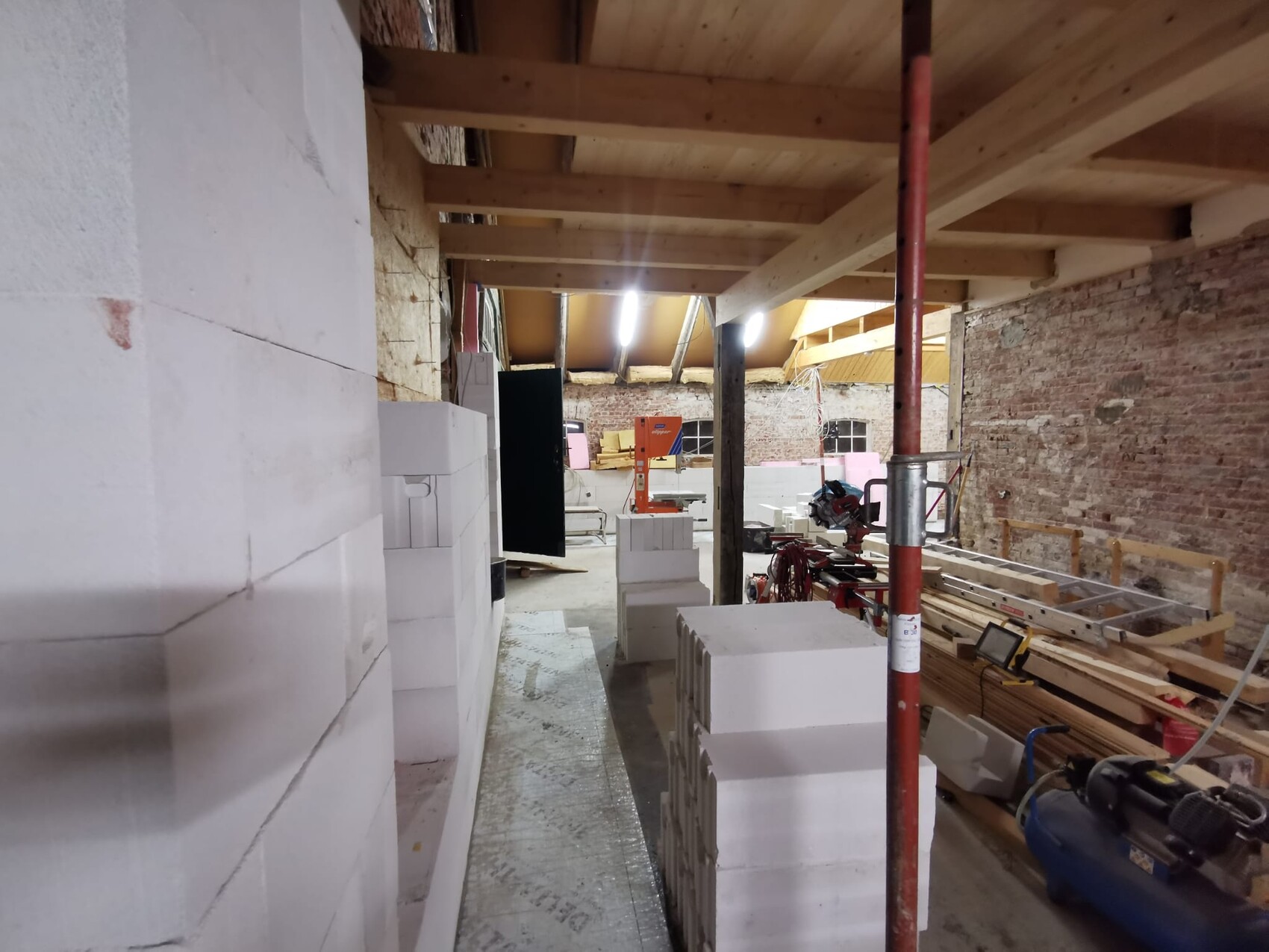 Innenraum-Baustelle mit gestapelten Styroporblöcken, Holzbalkendecke und Bauwerkzeugen.