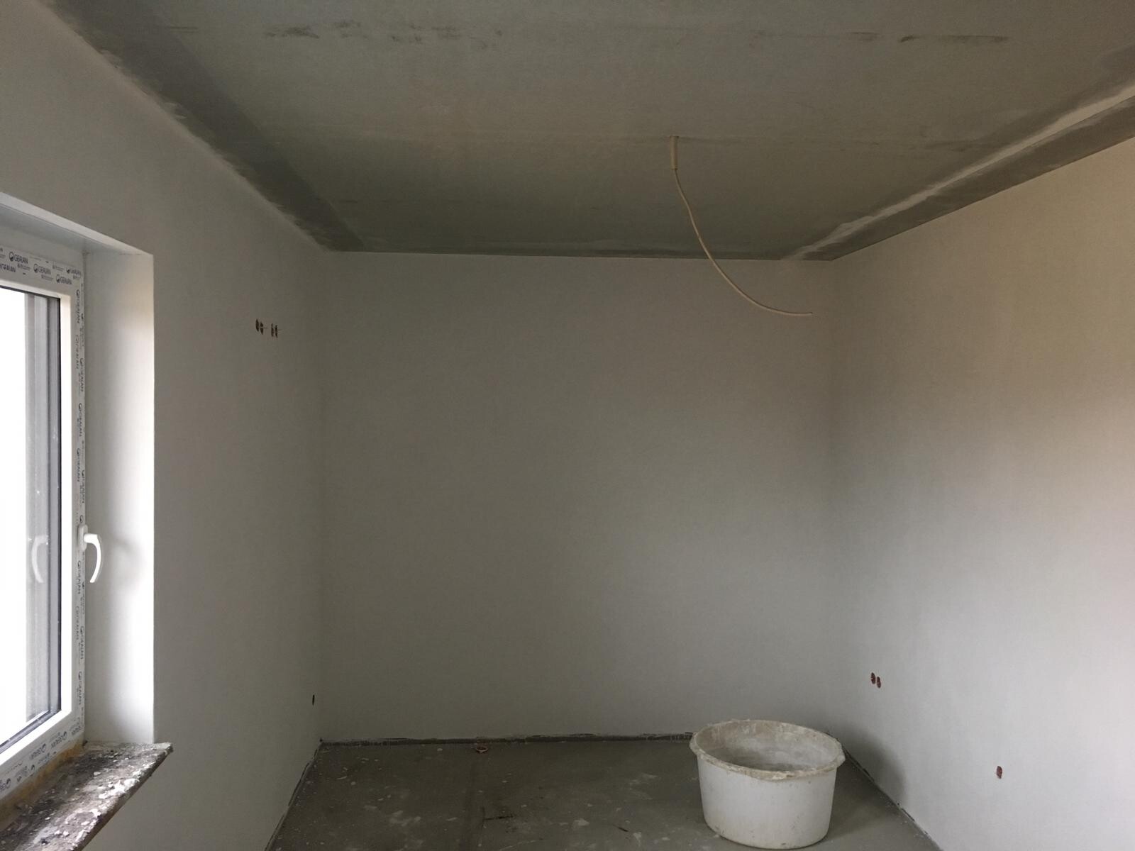 Unfertiger Renovierungsraum mit Fenster links, lose Kabeln von der Decke und Eimer auf dem Boden.