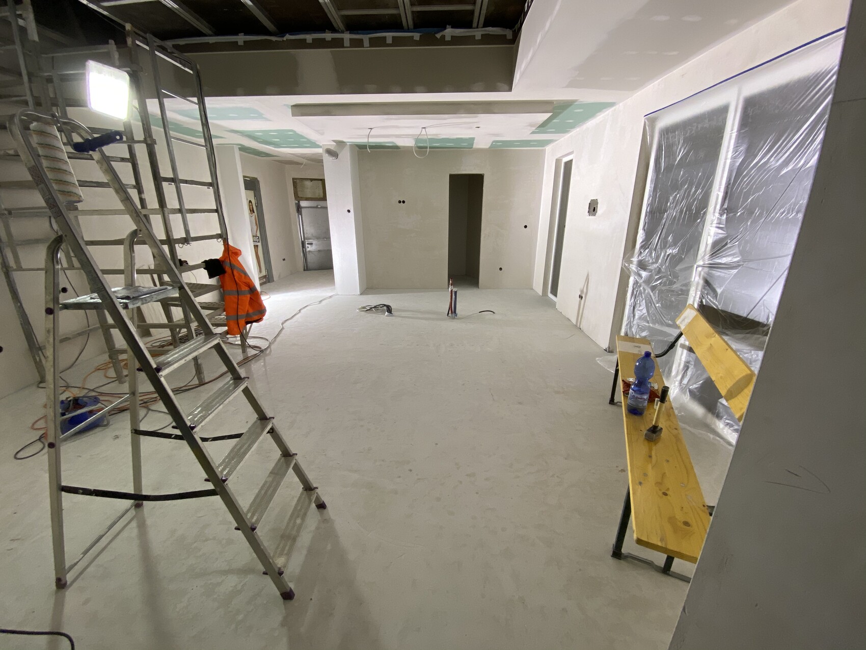 Innenraum einer Baustelle mit Gerüst, Leiter, orange Sicherheitsjacke, Werkbank mit Flasche, Plastikfolien an Türen.