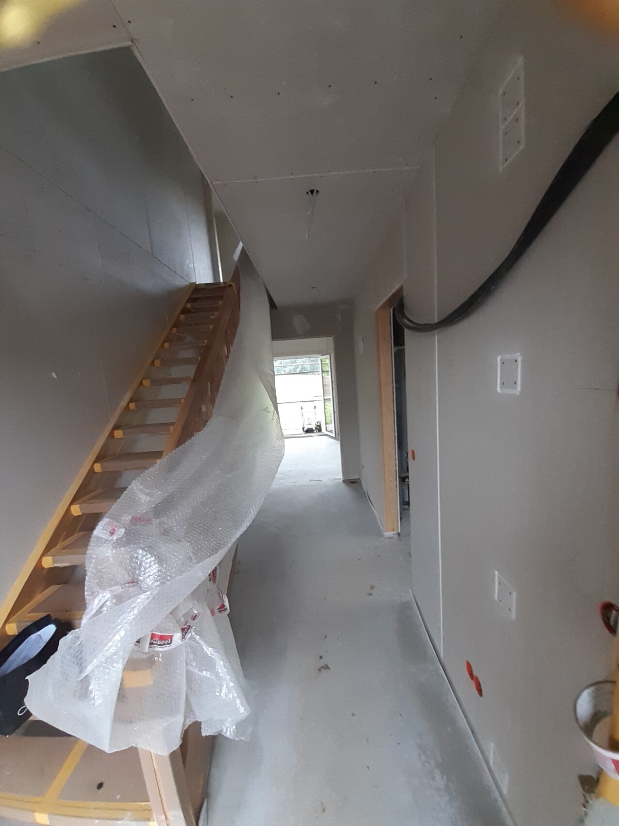Durchgang in einem unfertigen Hausflur mit Holztreppe links, Kabelkanal rechts, Baufolie.