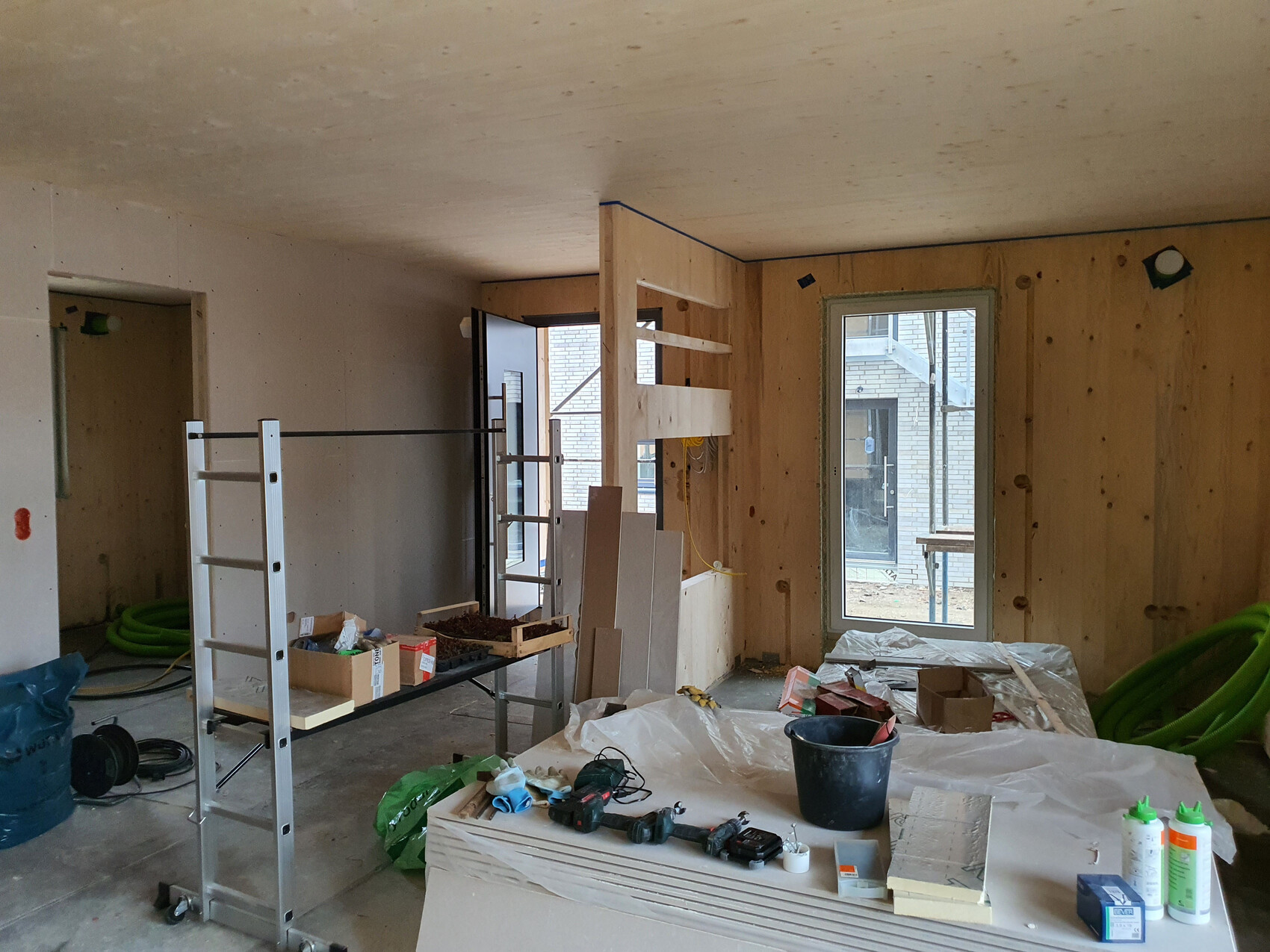 Renovierungsraum mit Gerüst, Holzplatten, Werkzeugen, Kartons und großer Fensterfront im Hintergrund