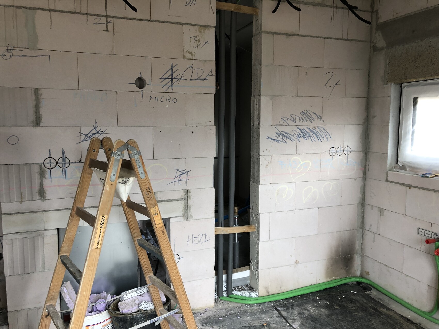 Baustelle: unfertige Blockwand, Holzleiter, grüner Schlauch am Boden, Graffiti, Fenster rechts.