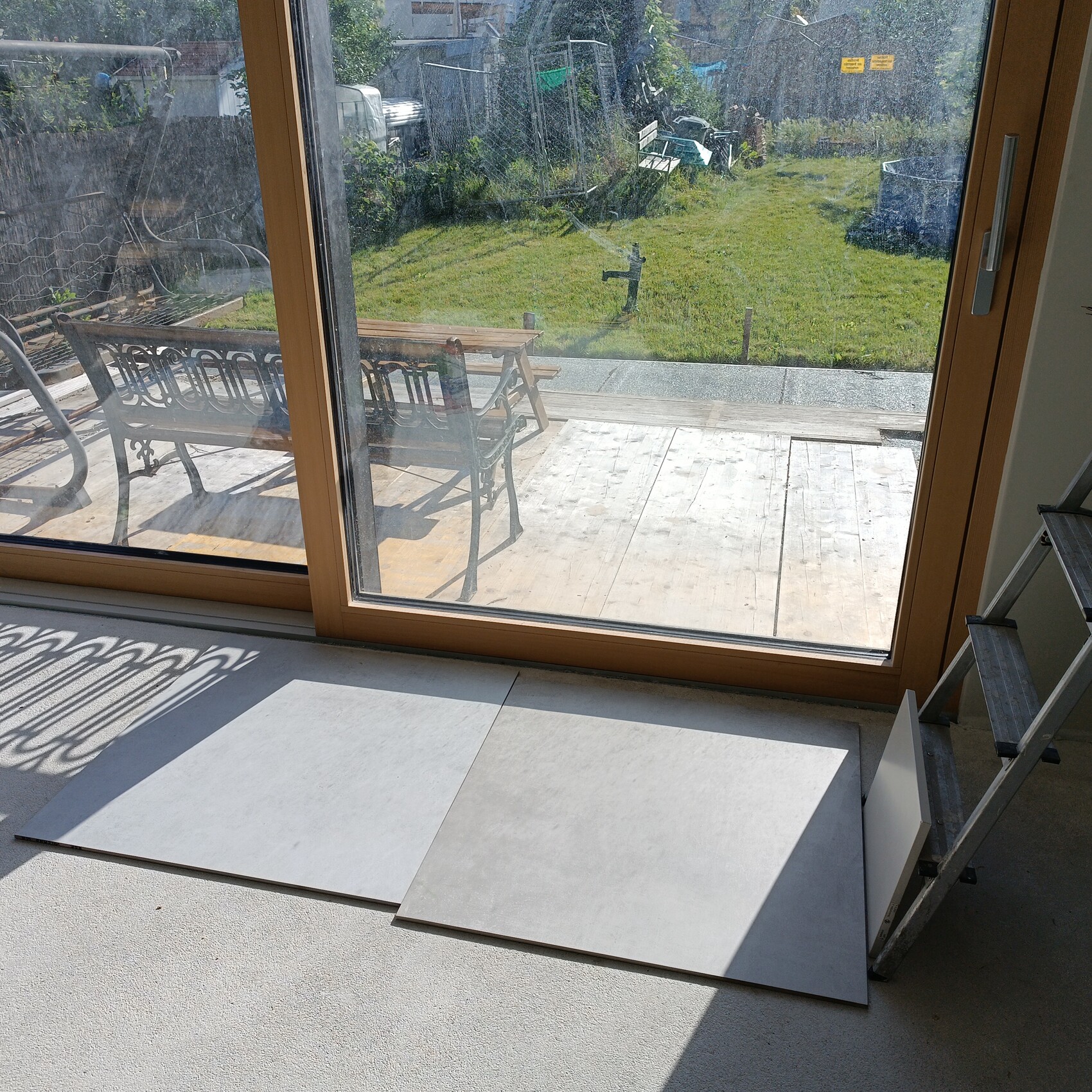 Blick durch eine Holz-Glas-Schiebetür auf eine Terrasse mit Metallstühlen vor grünem Garten.