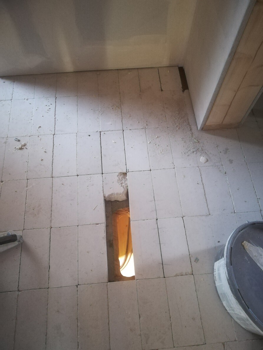 Teilweise entfernte Bodenfliesen; langes rechteckiges Loch zeigt orange Licht und Rohre darunter.