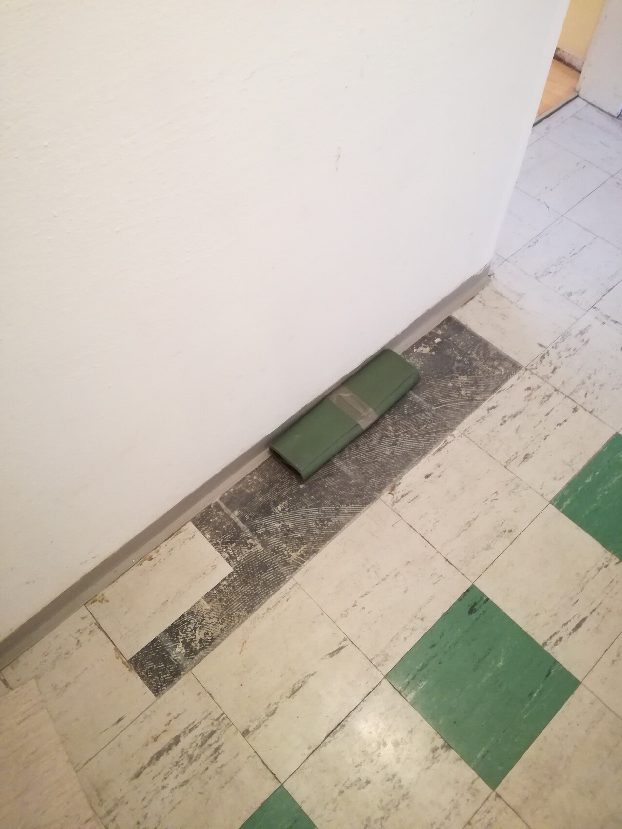 Grüner, länglicher Gegenstand liegt auf einem dunklen Bodenstreifen neben der weißen Wand.