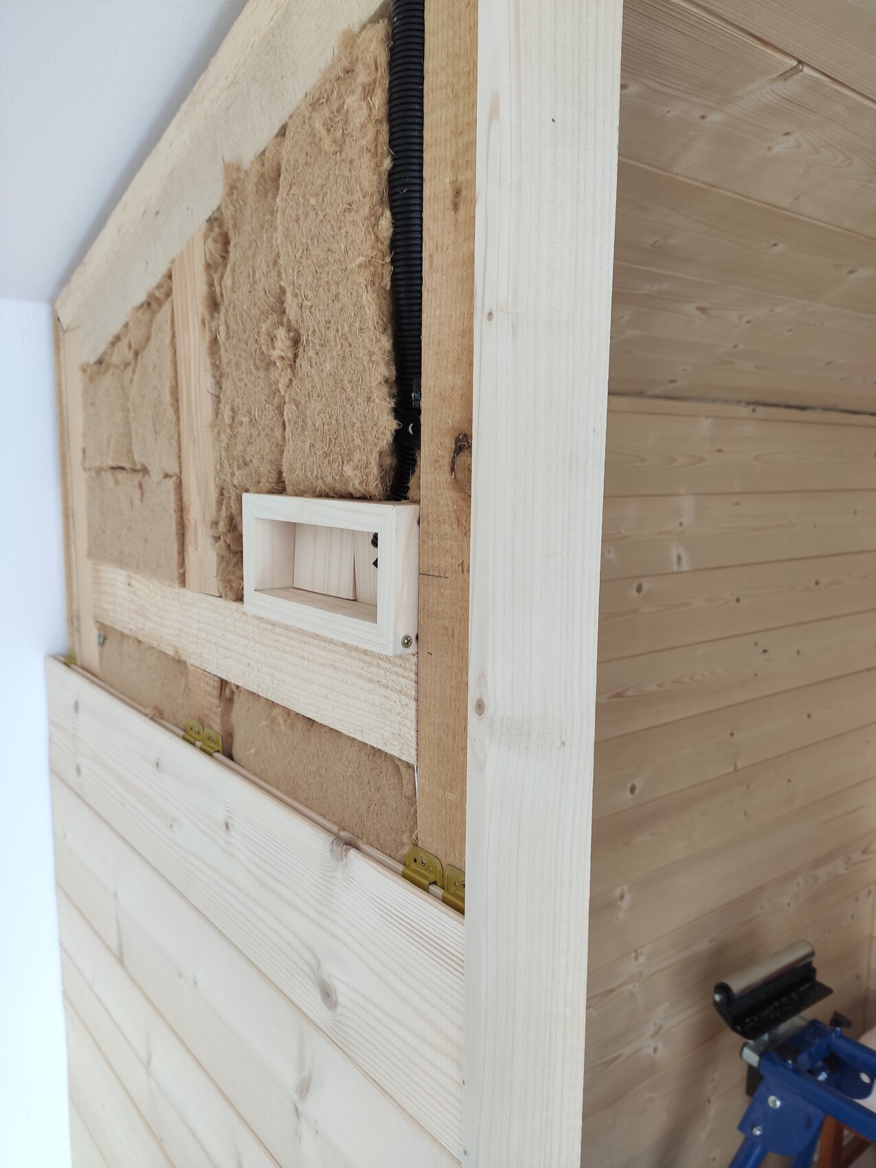 Holzinnenwand mit Dämmung, schwarzem Kabelkanal und einer kleinen rechteckigen Holzkiste.