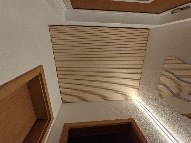 Innenraum: Decke mit holzfarbener Jalousie, Türrahmen links, wellenförmige Wanddeko rechts.