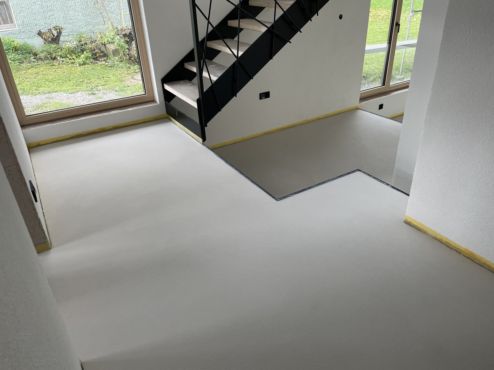 Neues Treppenhaus: grauer Epoxidboden, schwarze Metalltreppe, große Fenster, Gelb-Kantenschutz.