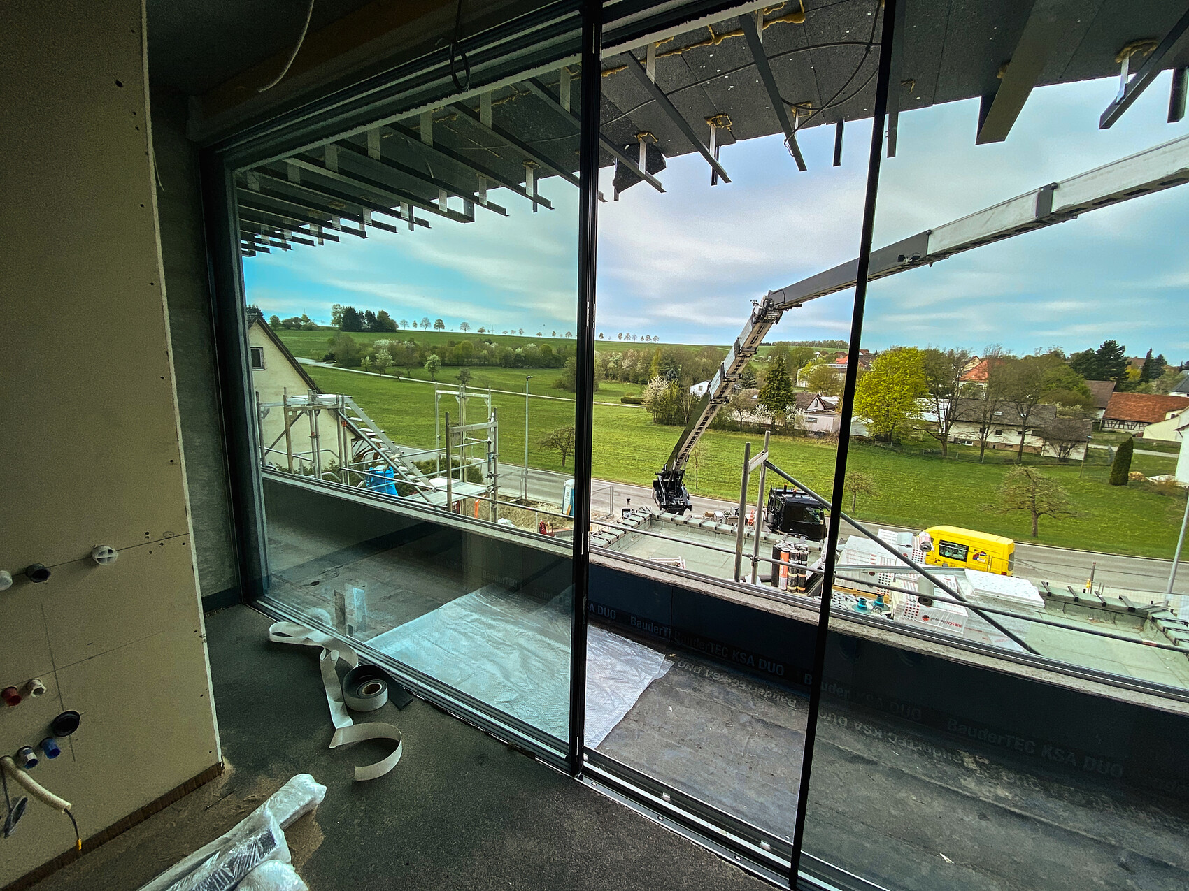 Blick durch Fenster auf Baustelle: Gerüst, Kranarm, Bauarbeiten; grüne Felder und Häuser.