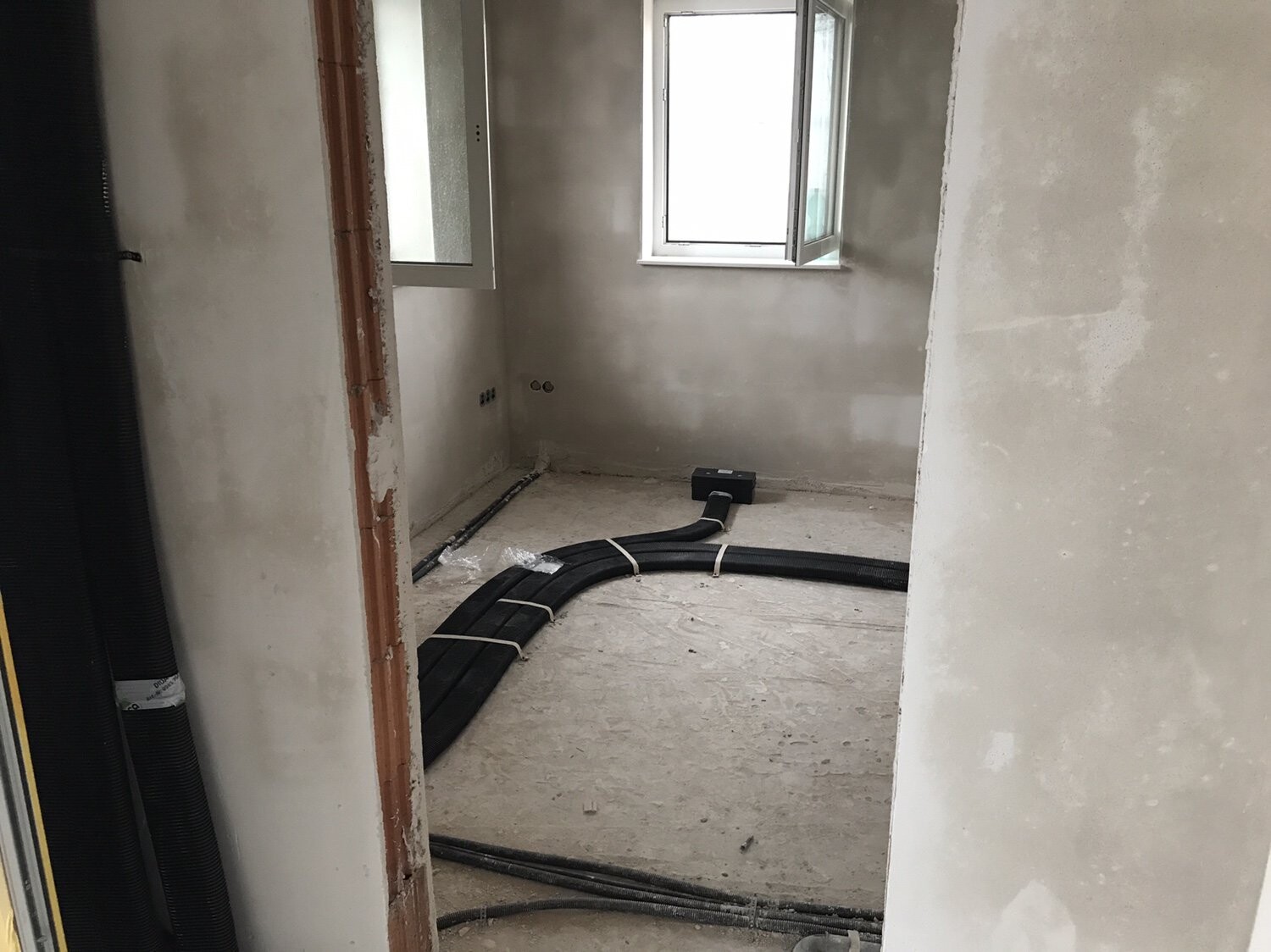 Baustellenzimmer mit offenem Fenster; schwarze Kabelrohre liegen gebündelt auf dem Boden.