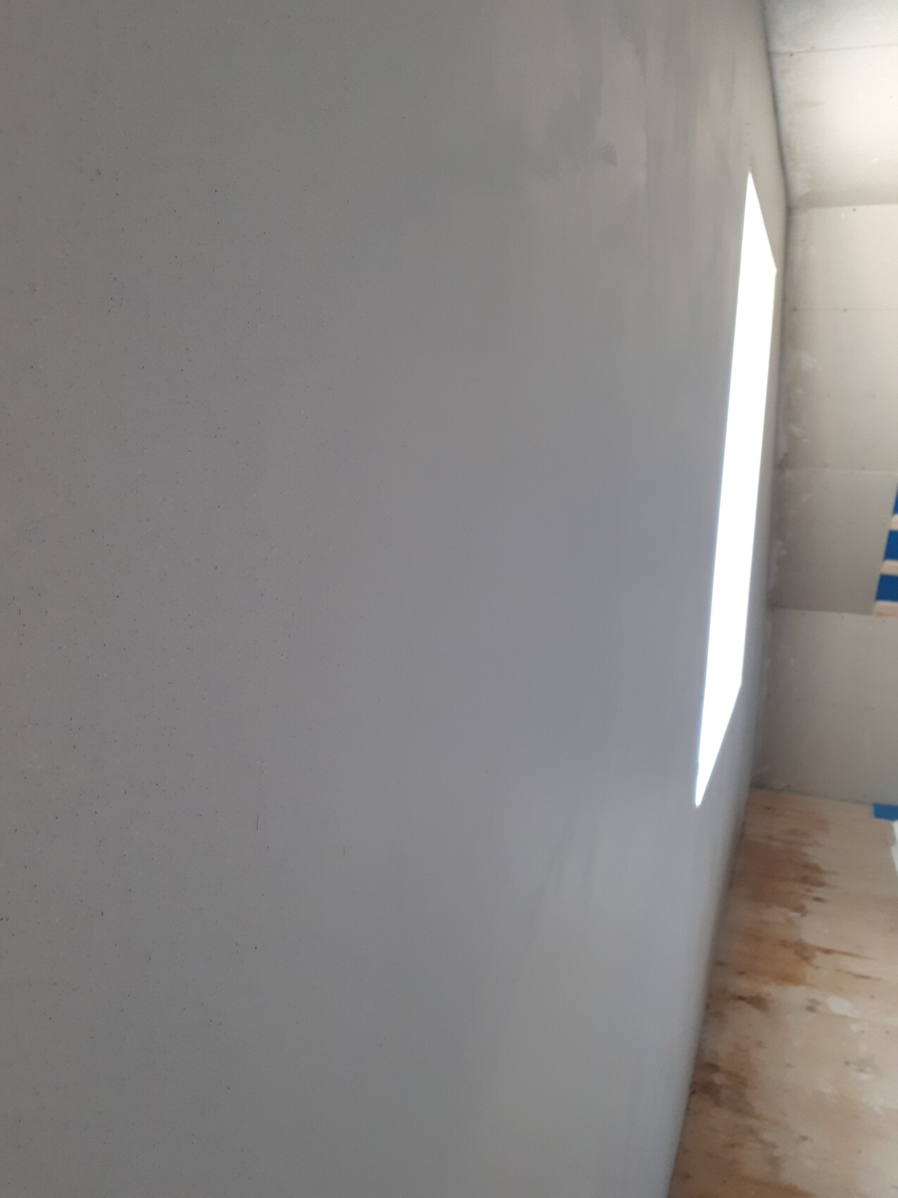 Graue Wand mit heller vertikaler Lichtöffnung rechts, Holzboden und blaues Malerband.