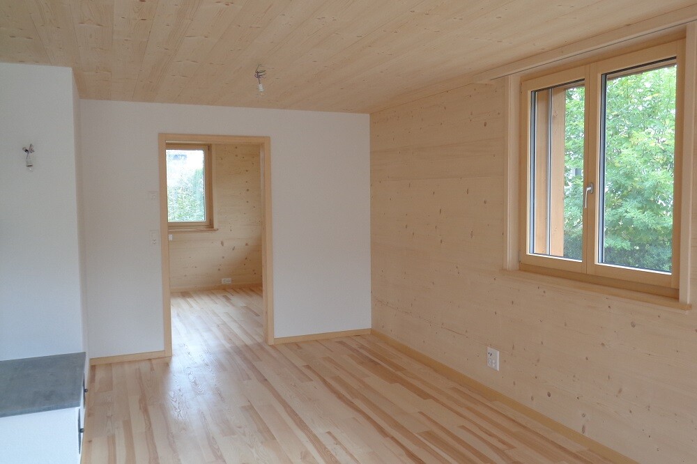 Heller Raum mit Holzwänden und Holzboden, großes Fenster rechts, Tür ins Nebenzimmer, unmöbliert.