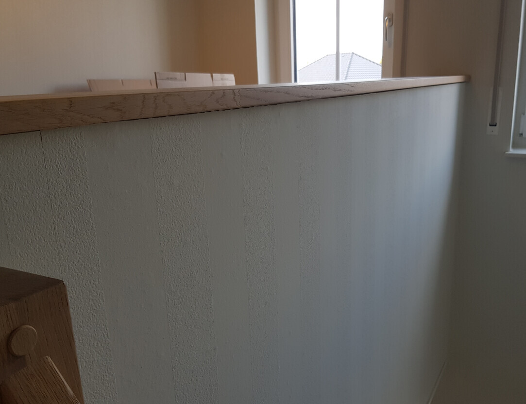 Heller Innenraum mit Holzgeländer über einer halben Wand; Fenster im Hintergrund.