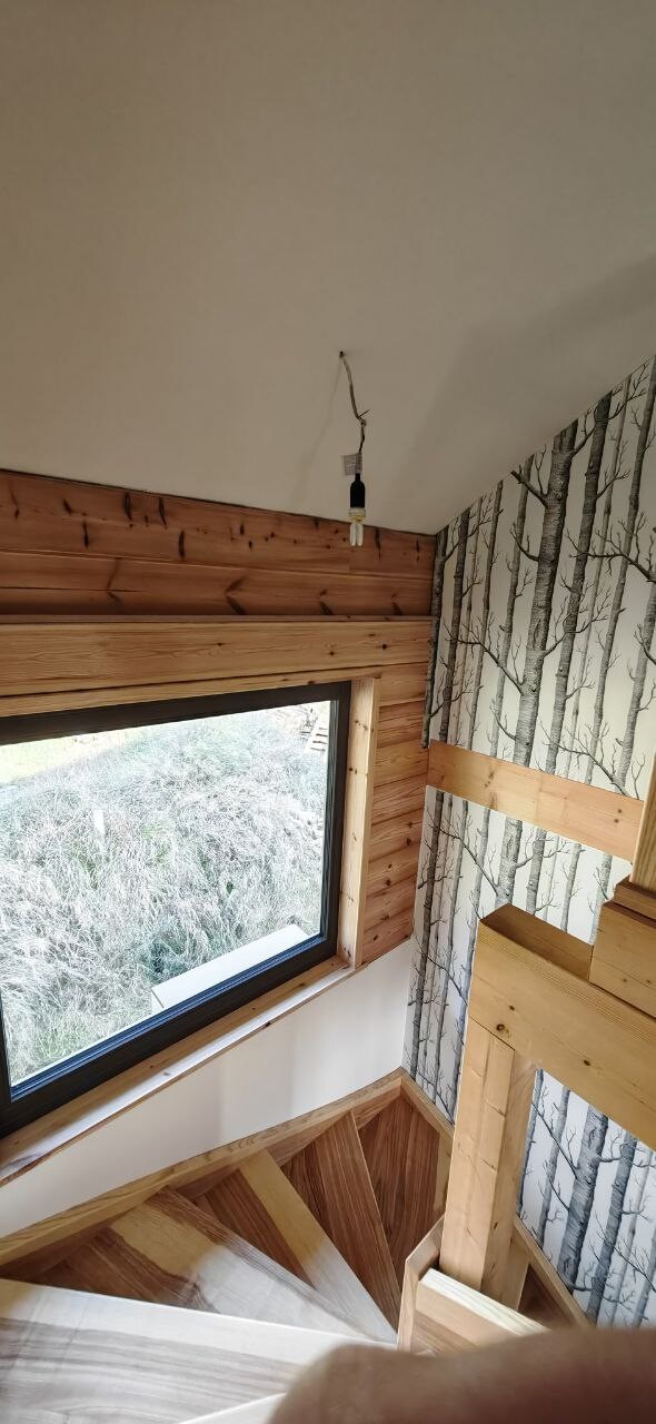 Holztreppe in hellem Innenraum, großes Fenster, Baumrinden-Tapete, lose hängende Leuchte.