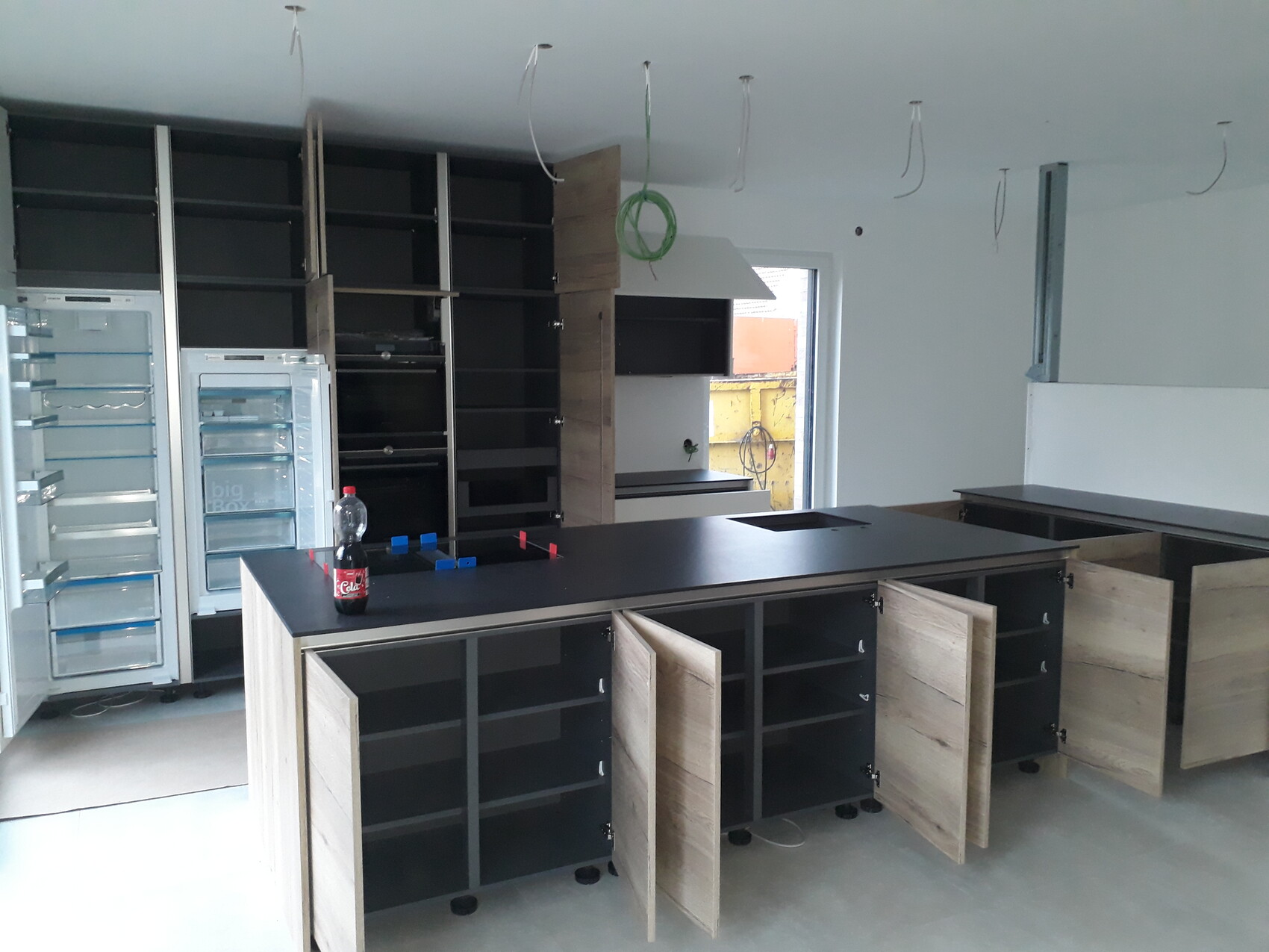 Küche im Bau mit dunklen Schränken, Insel, offenen Türen, leeren Kühlschrank und hängenden Kabeln.