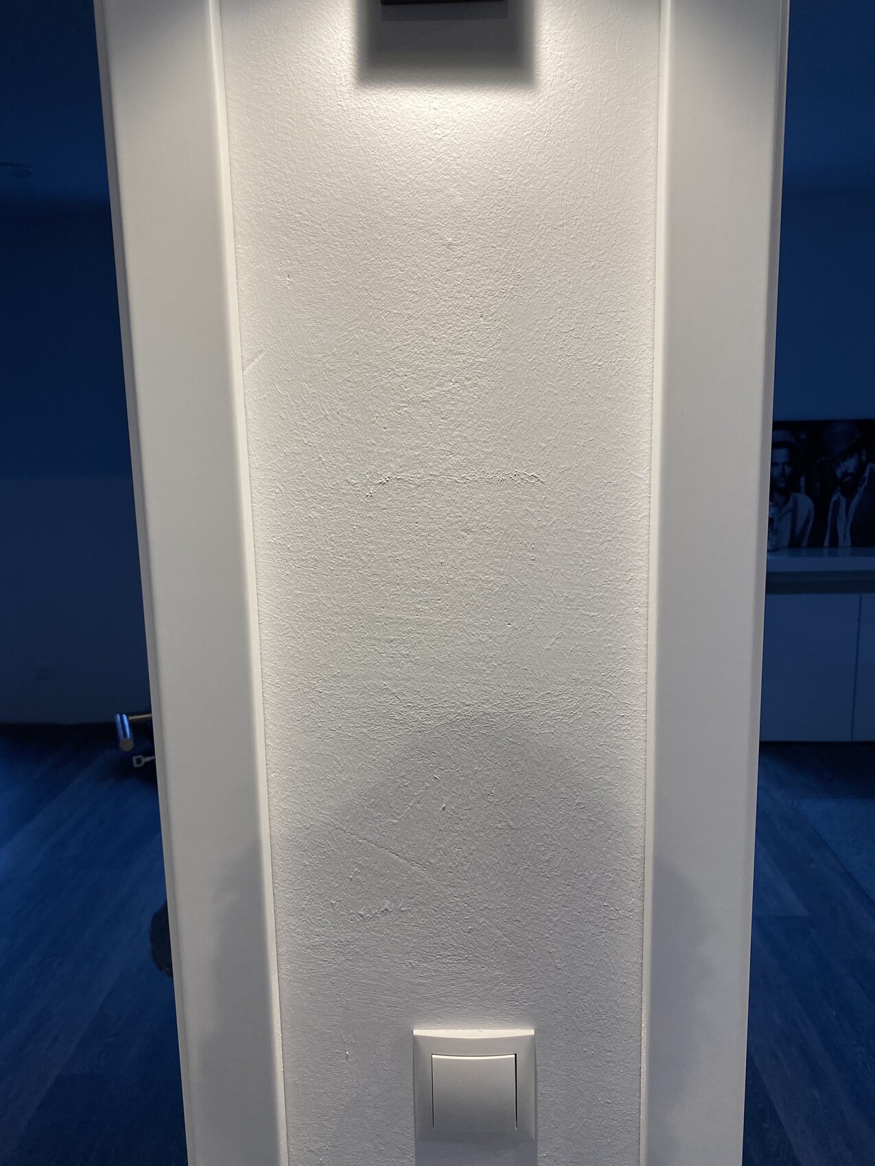 Weiße, texturierte Wand mit seitlichen weißen Rahmen; unten Lichtschalter, oben kleines dunkles Bauteil.