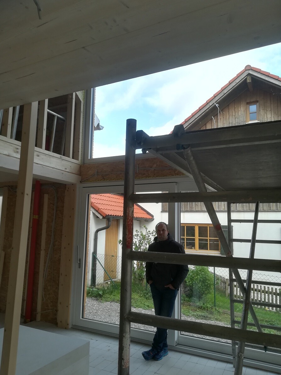 Mann steht in einem Neubau-Innenraum hinter Gerüst, große Glasfront, Blick nach draußen.