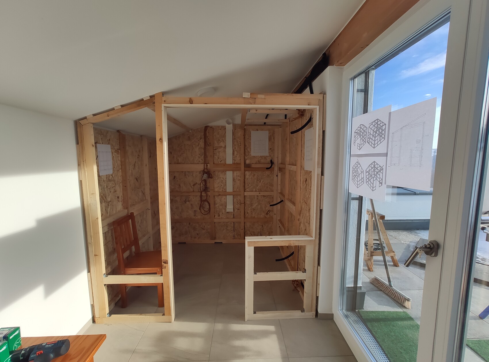 Kleiner Innenraum mit Holzkonstruktion, Holzstuhl, Werkzeugen und Skizzen an Glasfront.