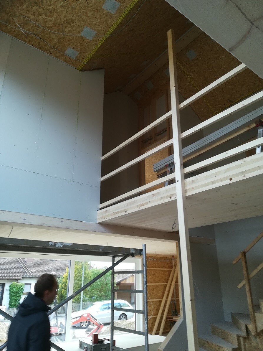 Neubau-Rohbau: Innenraum mit Holzgalerie, Geländer, Treppe und Bauarbeiter im Vordergrund.